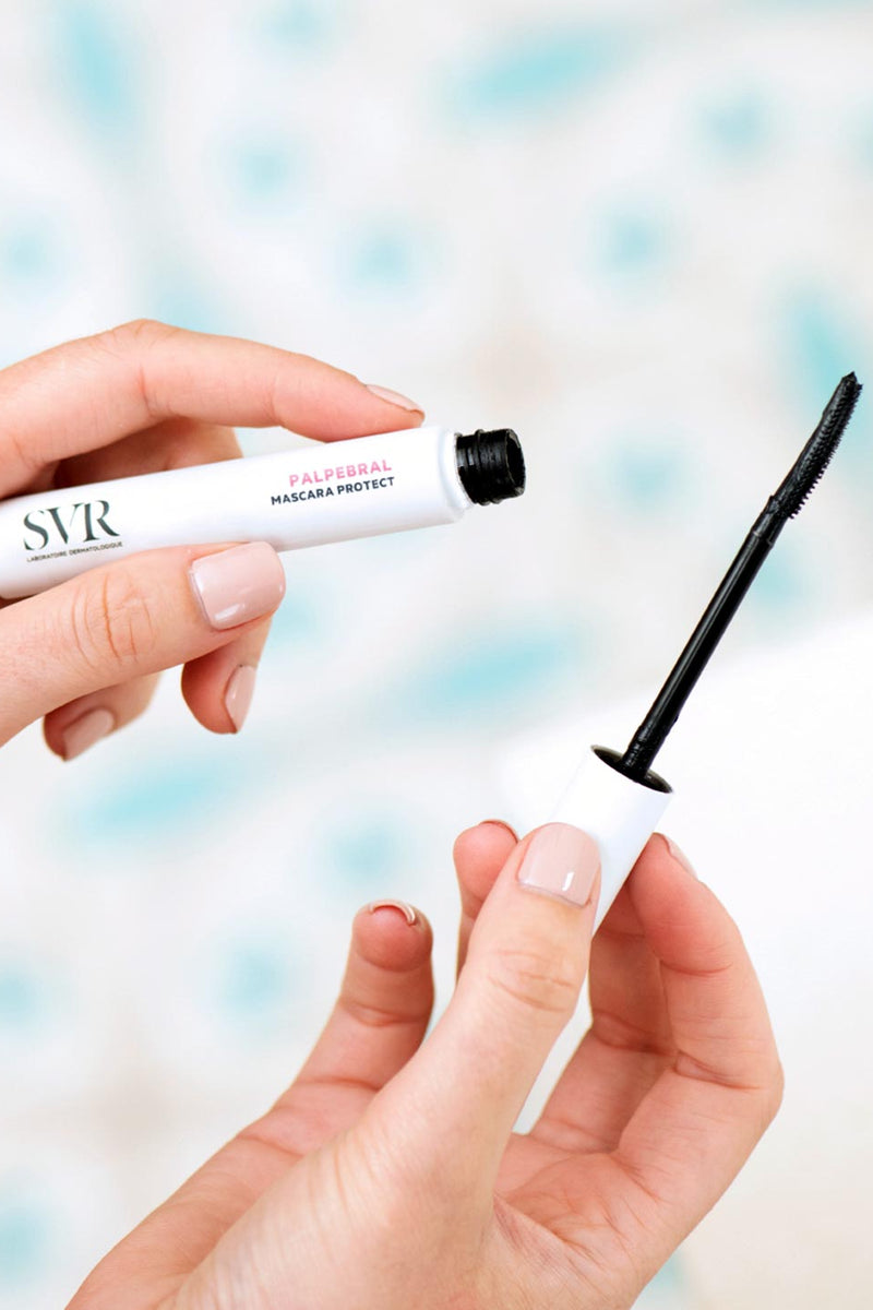 Mani che tengono il mascara SVR Palpebral Protect bianco con applicatore nero sottile e curvo su sfondo sfocato.