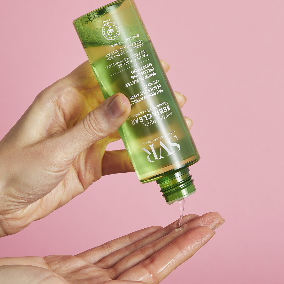 Flacone verde trasparente di SVR Sebiaclear Micro-Peel che versa l'essenza liquida sul palmo della mano su sfondo rosa.