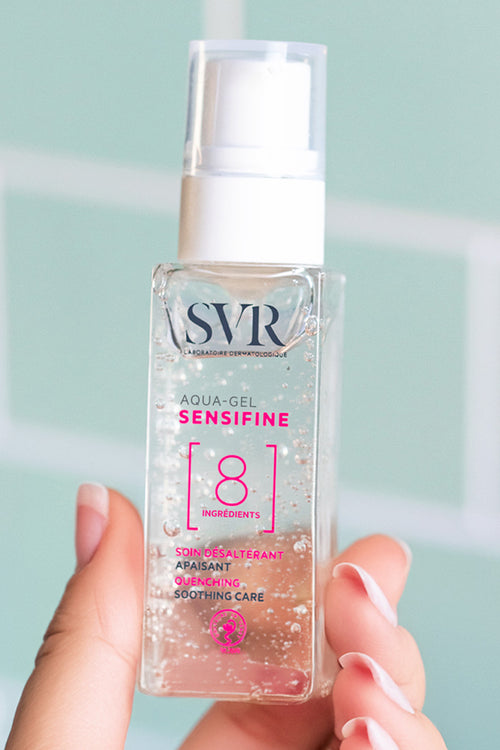 Flacone trasparente di SVR Sensifine Aqua-Gel con erogatore bianco, tenuto in mano su sfondo piastrellato color acqua.