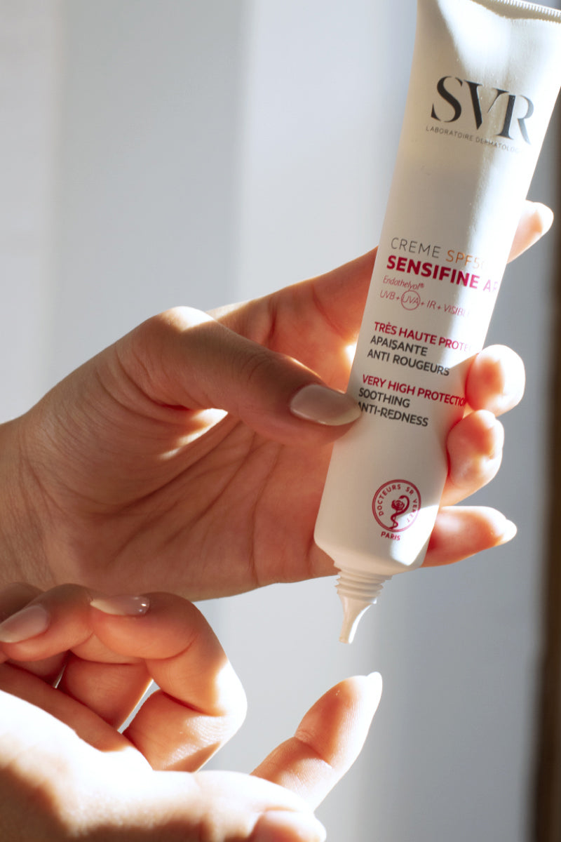 Mano che tiene un tubetto bianco di crema solare SVR Sensifine AR SPF50+ anti-rossore con scritte rosse e nere.