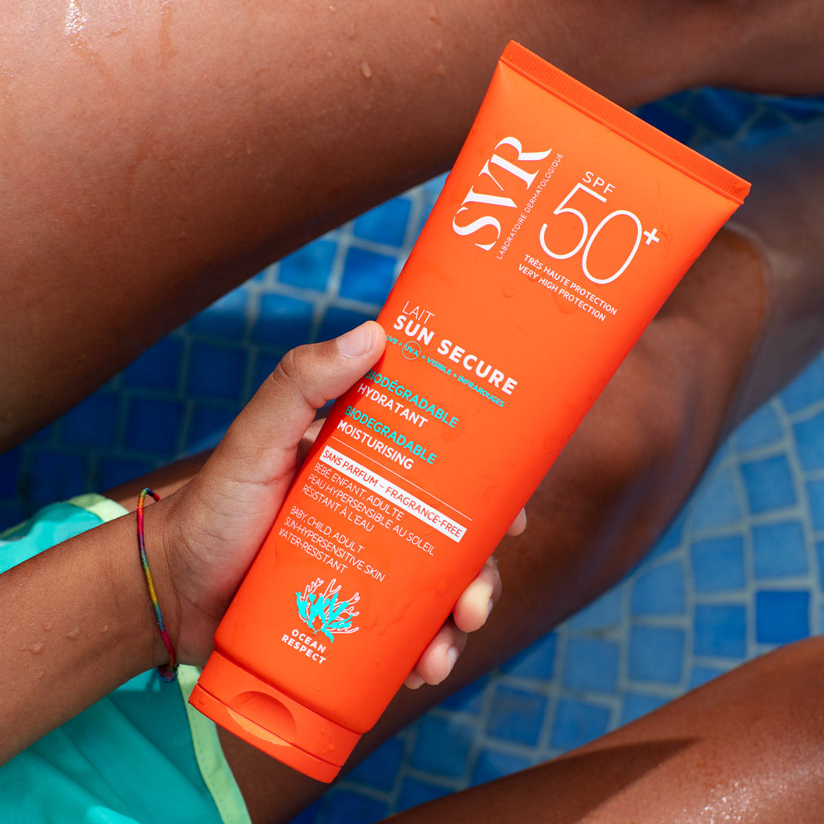 Tubo arancione di crema solare SVR Sun Secure SPF50+ tenuto in mano sopra una piscina piastrellata di blu.