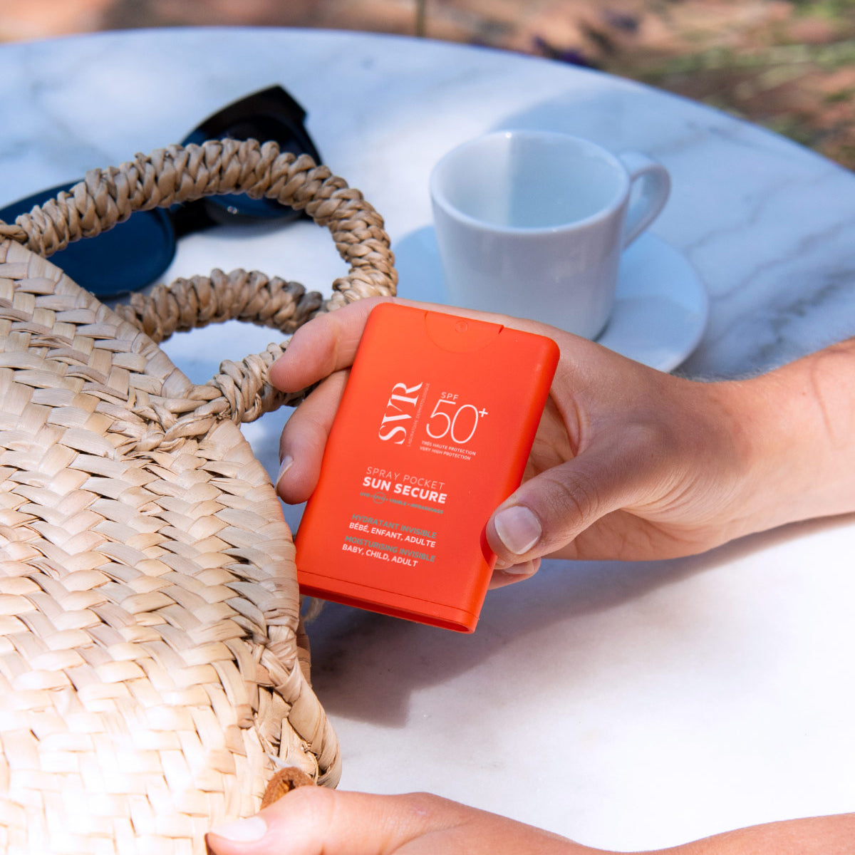 Flacone spray tascabile arancione SVR Sun Secure SPF50+ tenuto in mano vicino a una borsa di paglia su un tavolo.