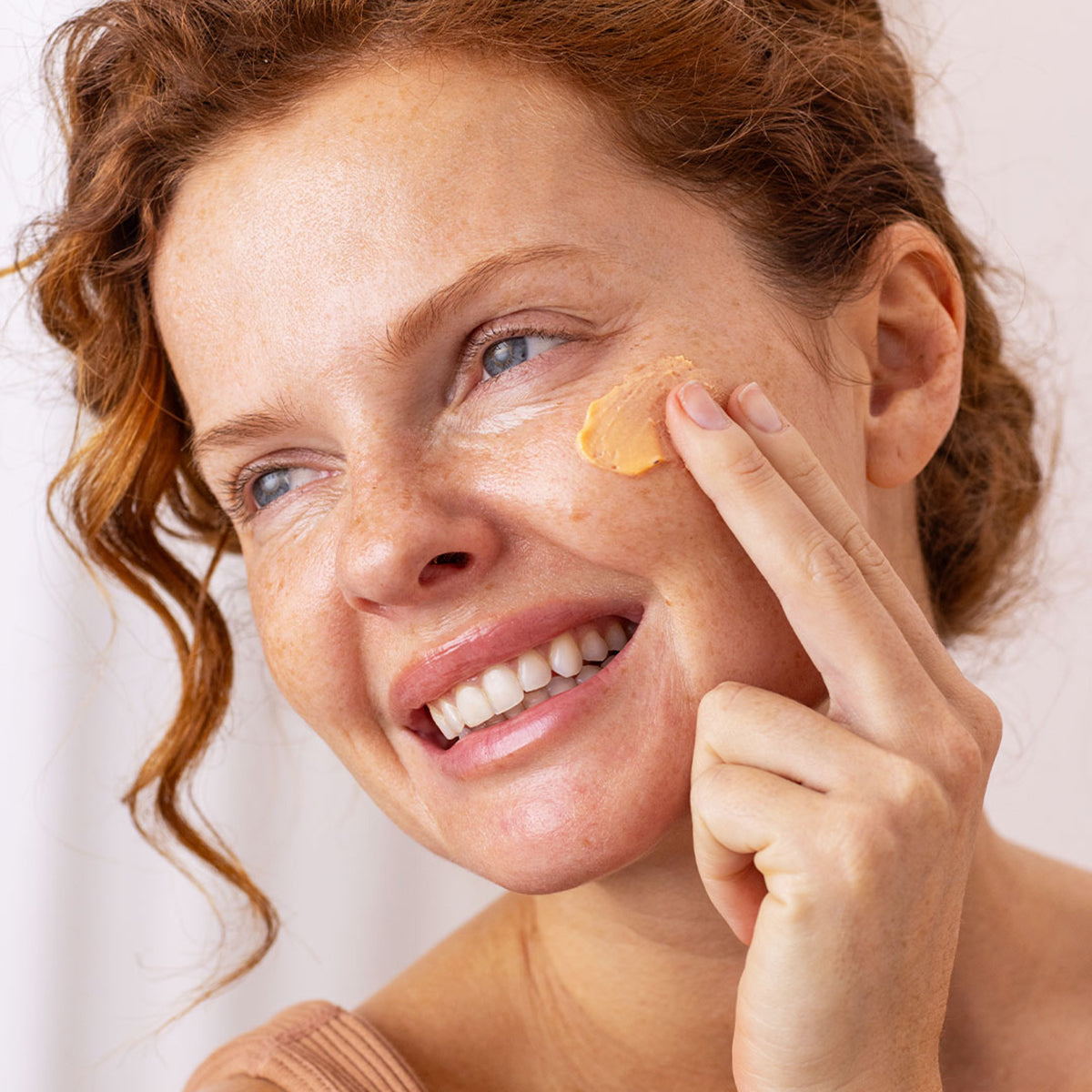 Donna con capelli rossi applica la crema solare color albicocca SUN SECURE Blur SPF50+ sulla guancia.