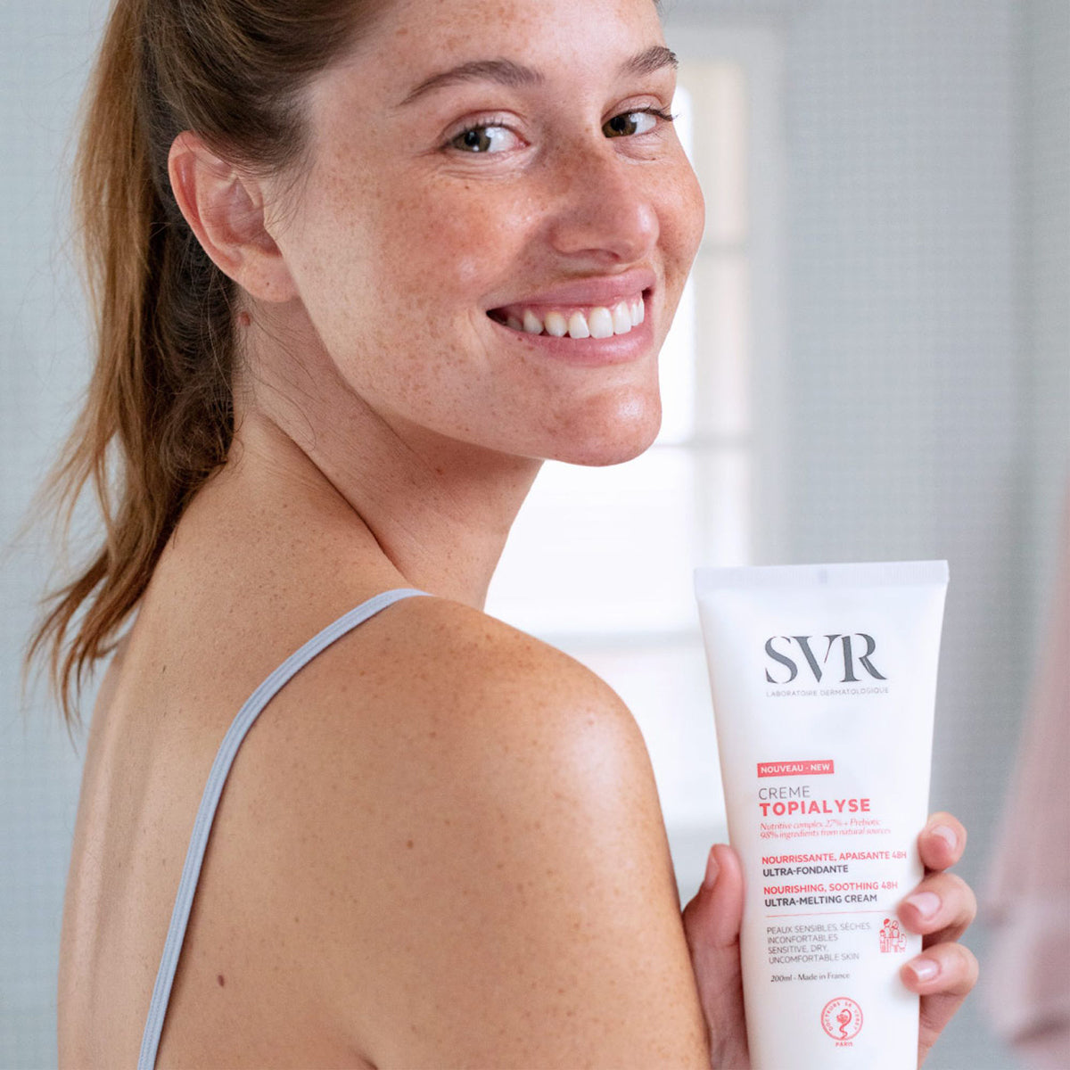 Donna sorridente tiene in mano il tubo bianco di crema idratante SVR TOPIALYSE da 200ml per pelli secche e sensibili.