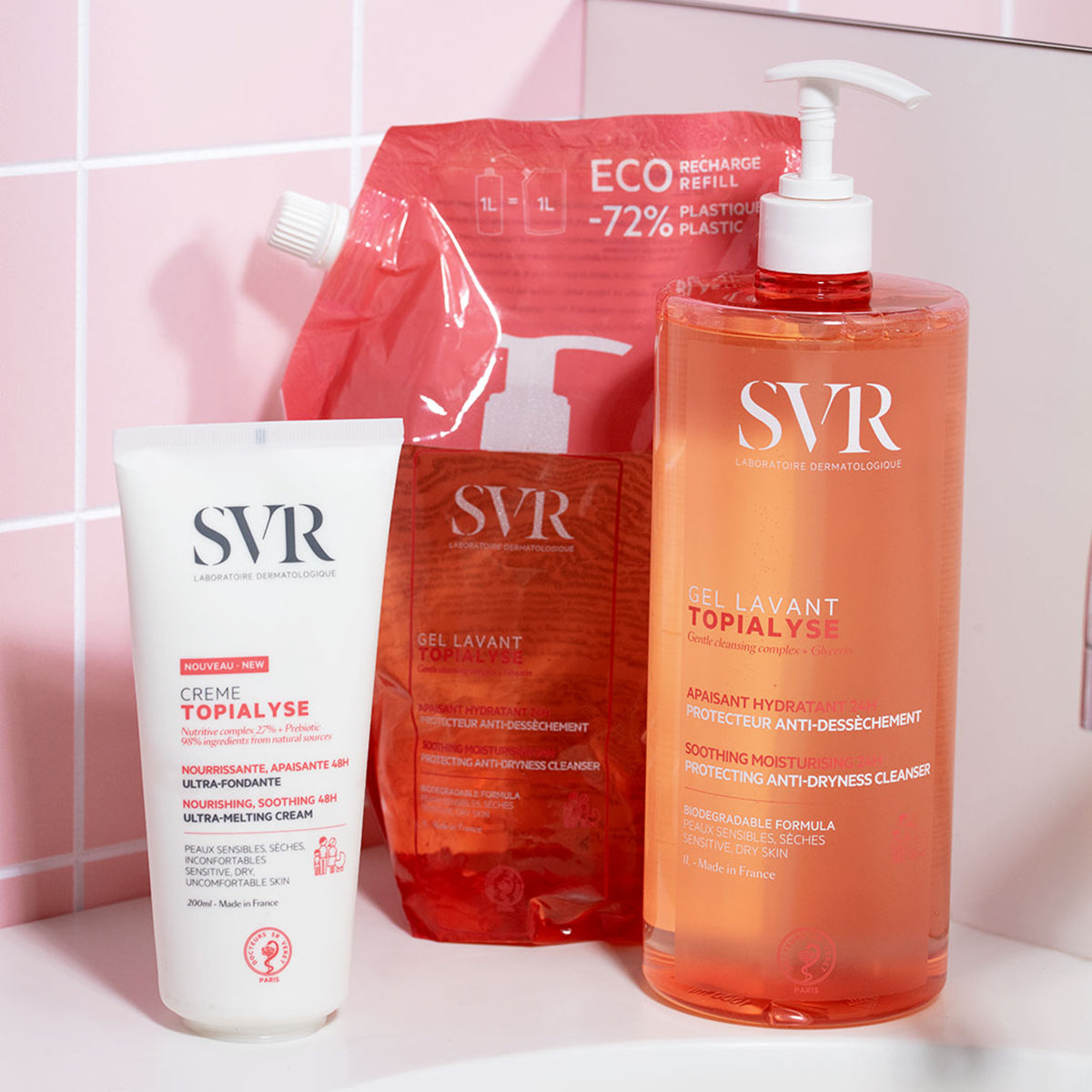 Prodotti SVR Topialyse: crema in tubo bianco, ricarica eco e flacone da 1L di gel lavante arancione su sfondo piastrellato rosa.
