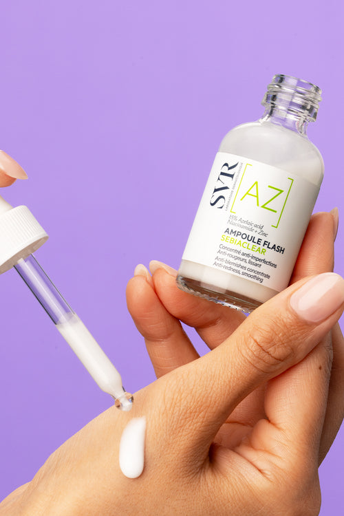 Flacone di SVR Sebiaclear Ampoule Flash con contagocce che applica il siero bianco sulla mano su sfondo viola.