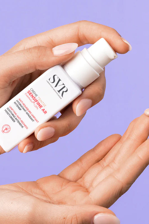 Flacone bianco di SVR Sensifine AR Crème Teintée con erogatore a pompa e goccia di crema colorata sul dito su sfondo lilla.