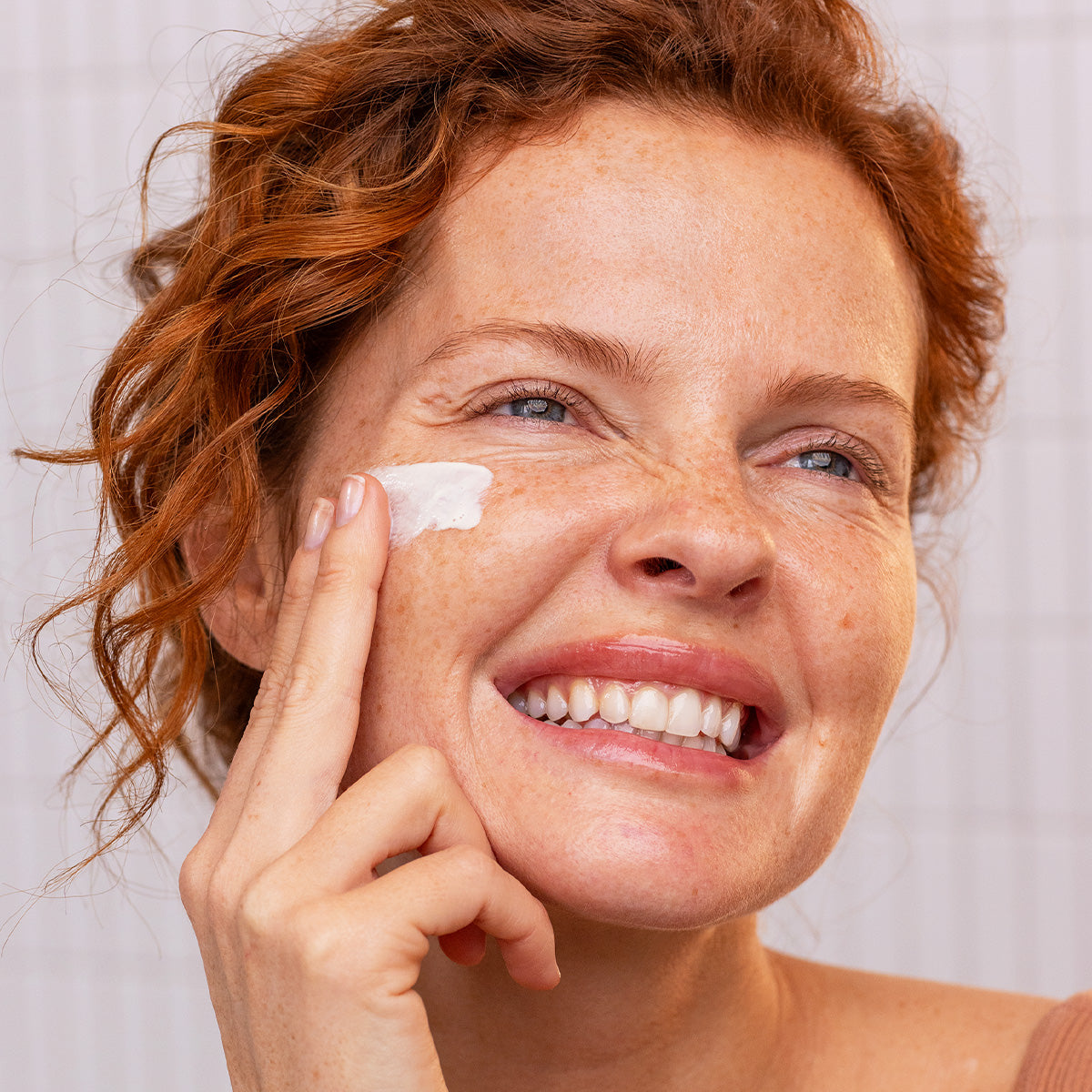 Donna sorridente con capelli rossi applica la crema solare Sun Secure Fluide SPF50+ sulla guancia in un ambiente luminoso.