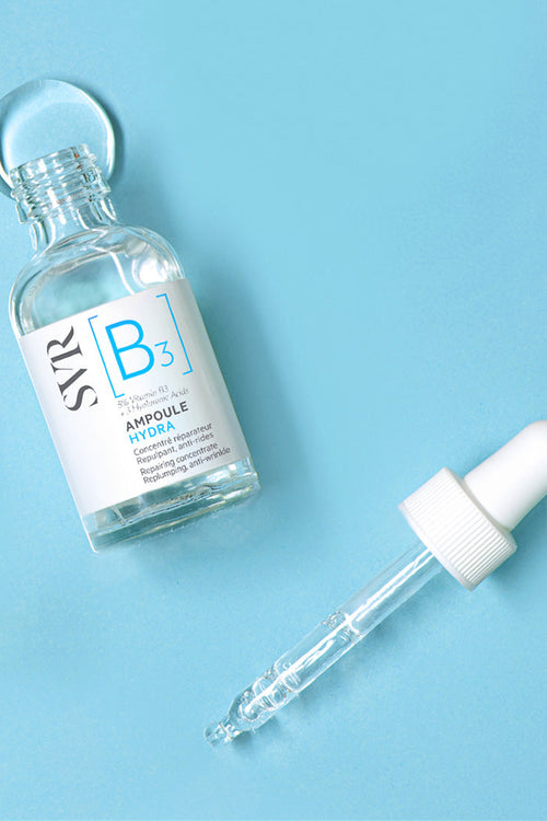 Flacone in vetro trasparente SVR Ampoule Hydra con contagocce separato su sfondo azzurro.
